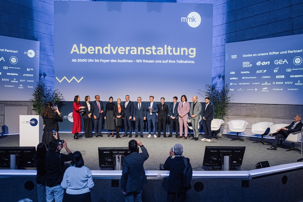 OLD text: A group of people stand on a stage at an evening event. In the background is a large banner with the inscription 
