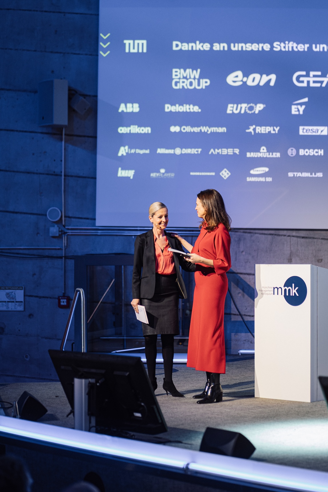 Zwei Frauen stehen auf einer Bühne vor einem Bildschirm mit Logos von Unternehmen. Eine Frau trägt ein rotes Kleid, die andere ein schwarzes Outfit.