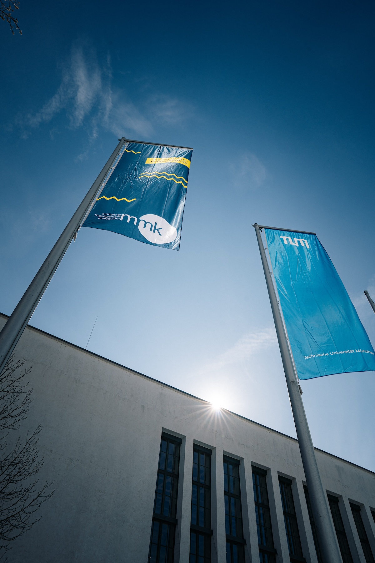 Two flags fly in front of a modern building, in clear skies and sunshine.